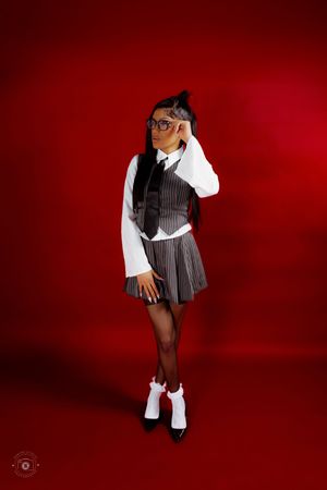 Young woman in school girl costume with vest, tie, plaid skirt, glasses, and white socks on red background.