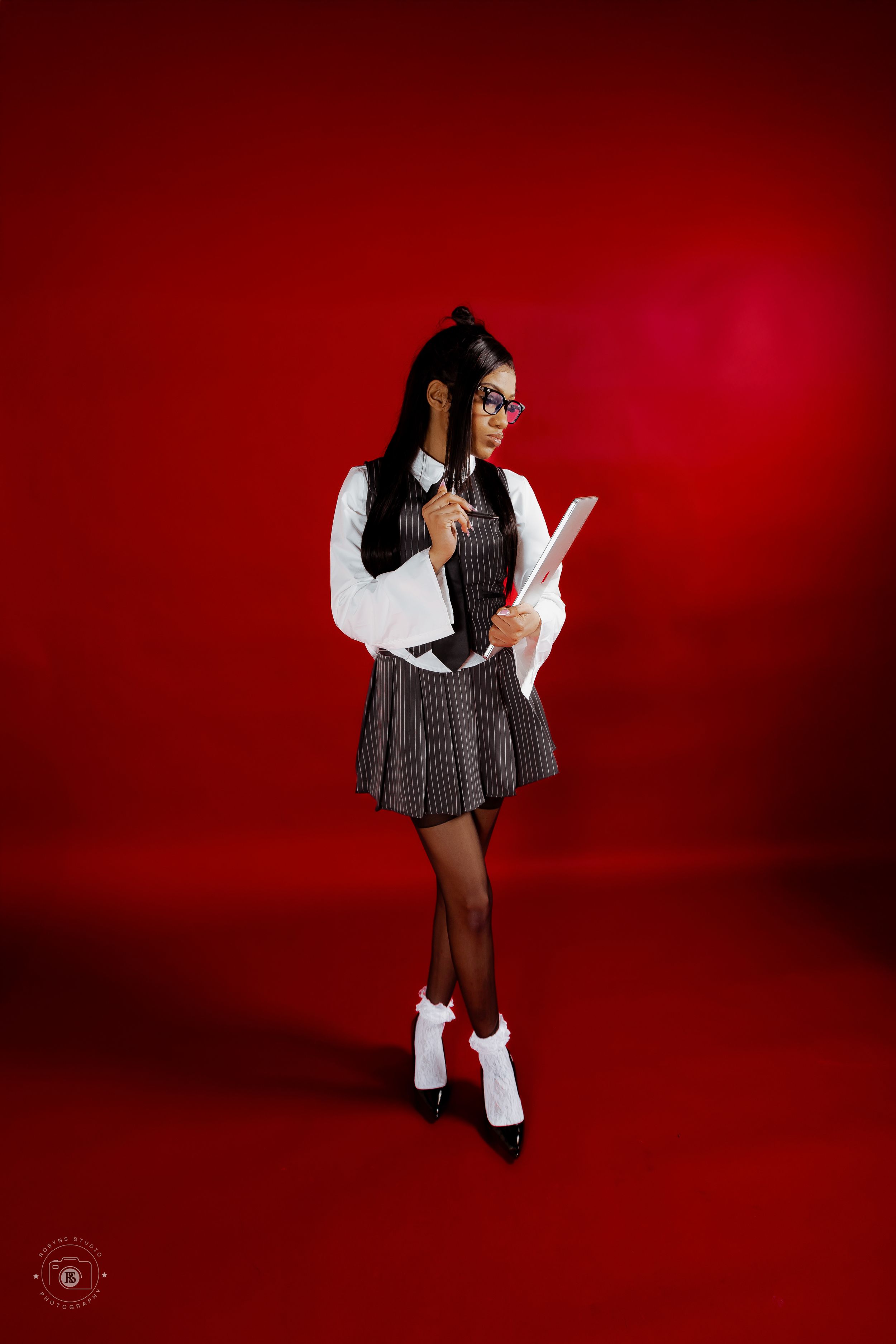 Young woman in school girl costume with glasses, pleated skirt, white blouse, and knee socks on red background.