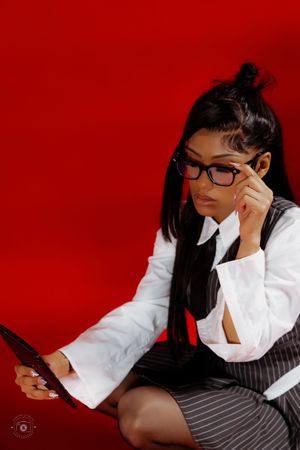 Woman in pinstripe skirt suit and glasses reads a tablet while sitting against a red background.