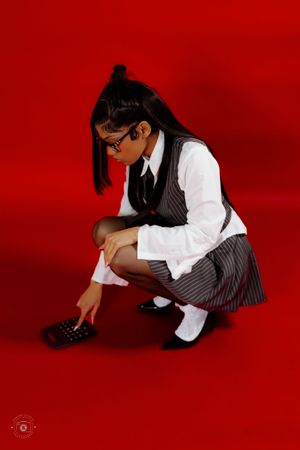 Young woman in school uniform with glasses crouching on red background, holding a calculator.