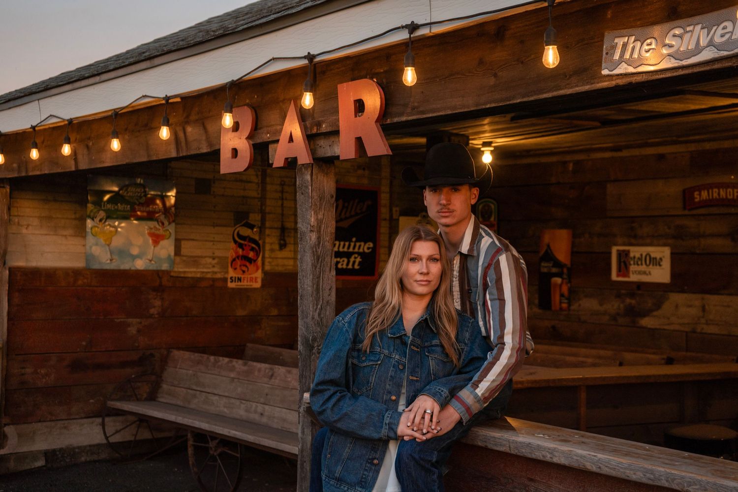 A couple poses in front of a rustic wooden bar at golden hour, man wearing cowboy hat and woman in denim jacket.