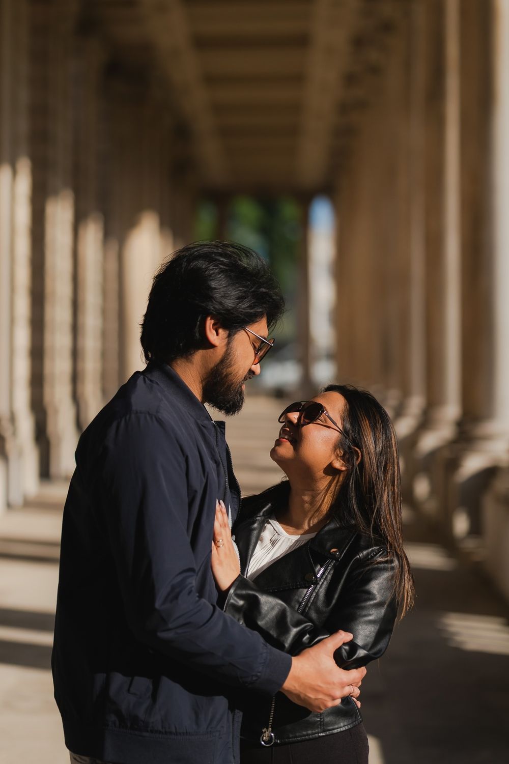 Romantic couple embracing between stone columns, woman in leather jacket looks up at bearded man in navy jacket.