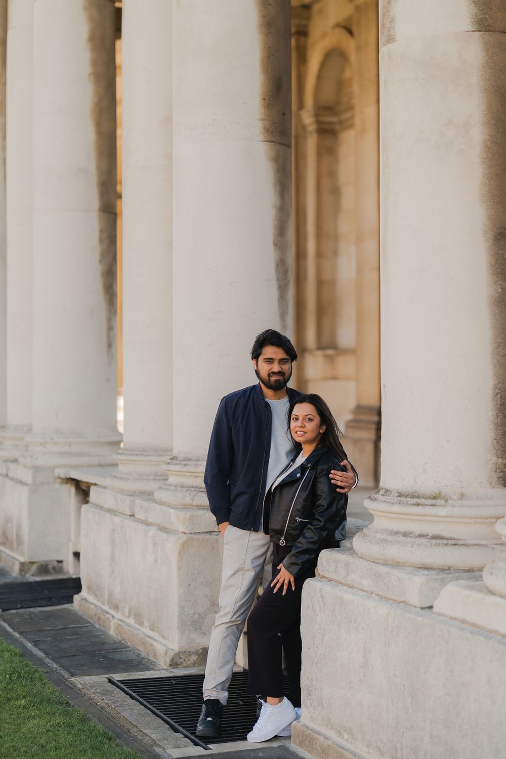 Couple posing in front of grand stone columns, man in navy blazer and woman in black leather jacket.