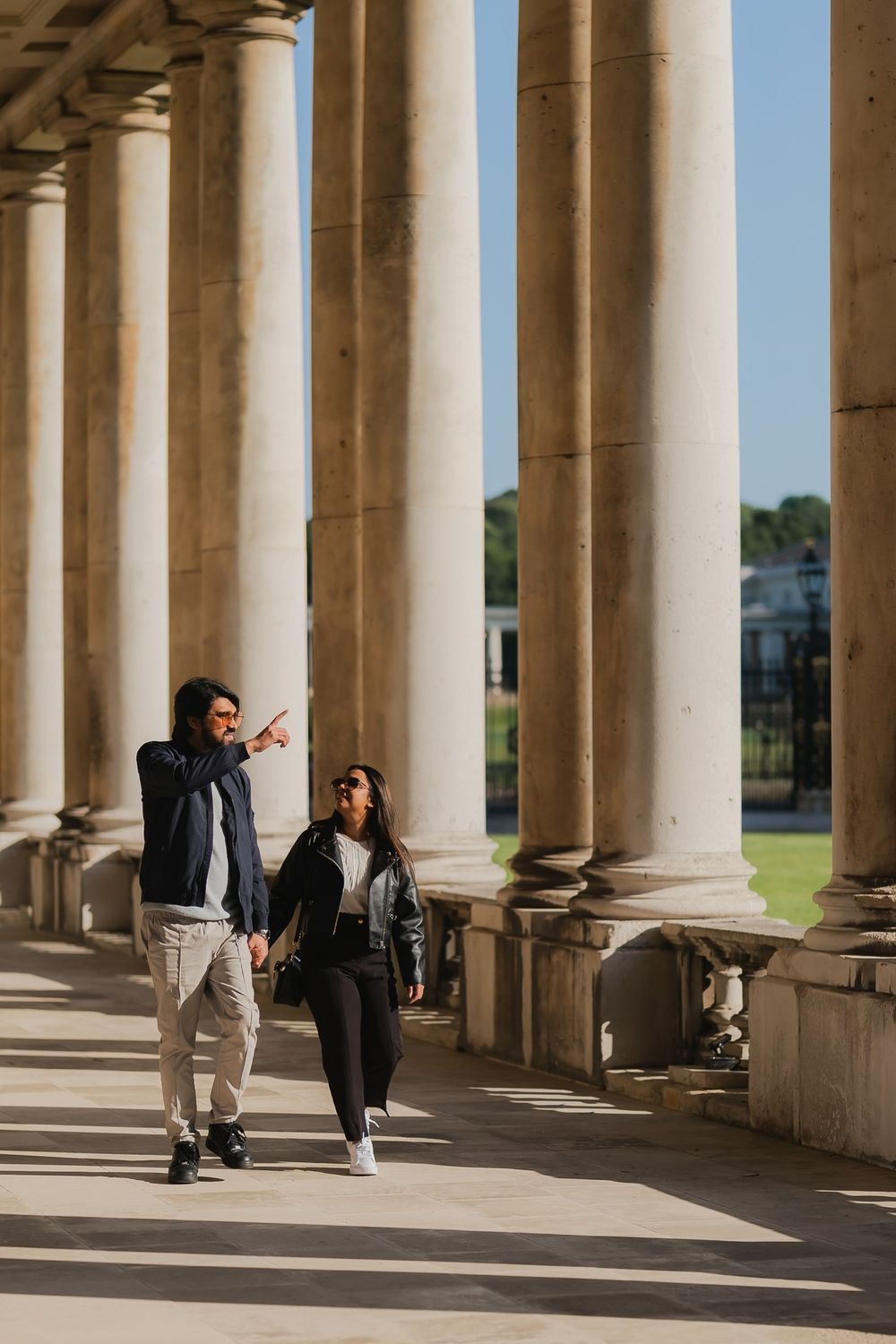 Couple walking hand in hand among grand stone columns of a classical building, man pointing into the distance.