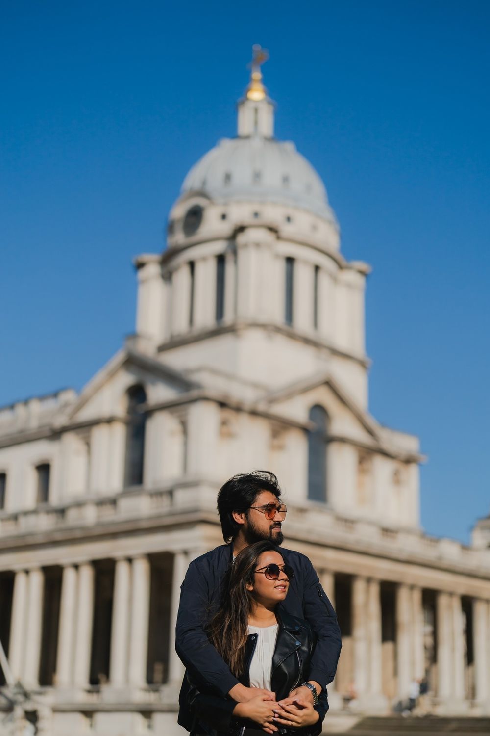 Couple embracing in front of the Old Royal Naval College dome in Greenwich, London, on a clear sunny day.