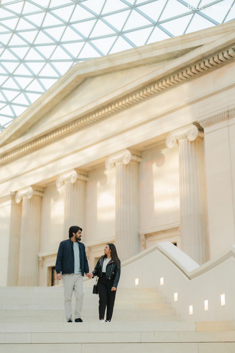 Couple holding hands on steps inside the British Museum, framed by grand ionic columns and glass ceiling.