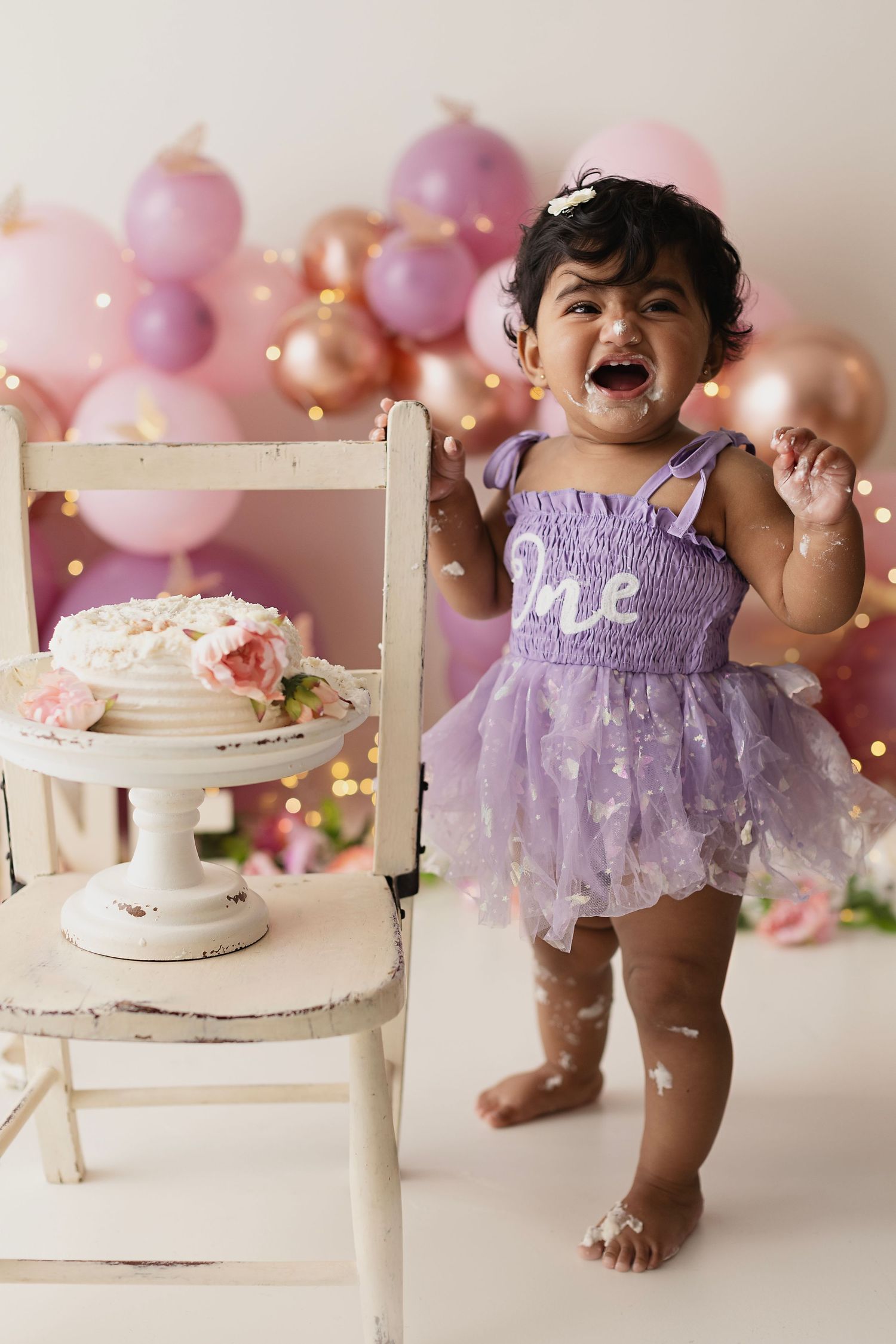 A joyful baby girl in a purple tutu dress celebrates her first birthday with a smash cake and pink balloon backdrop.