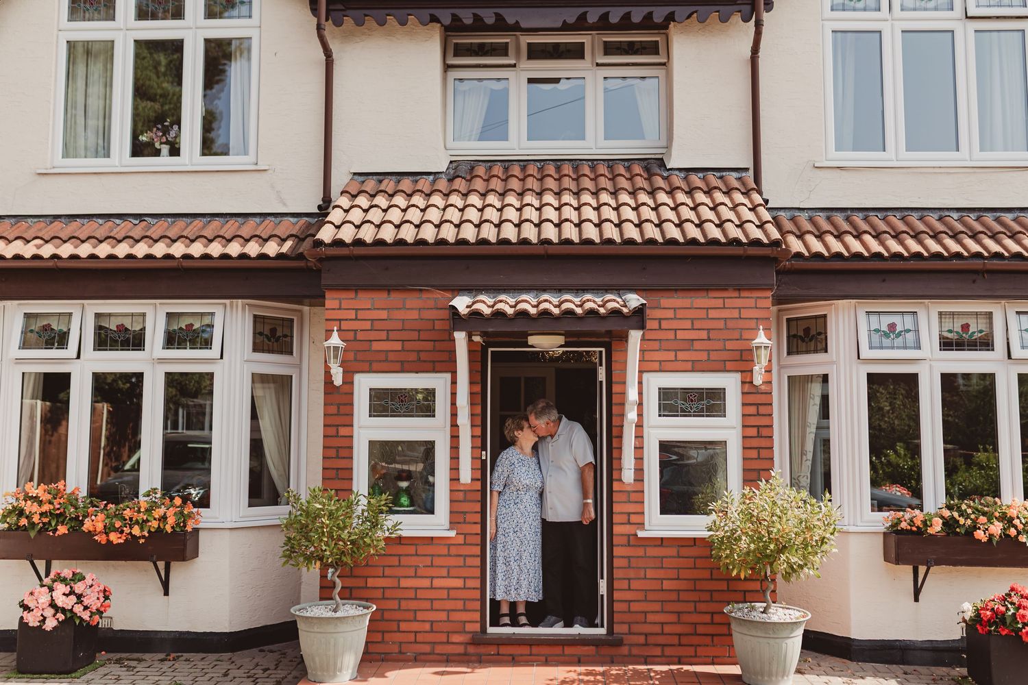 Elderly couple standing together in the doorway of a charming brick house with flower boxes.
