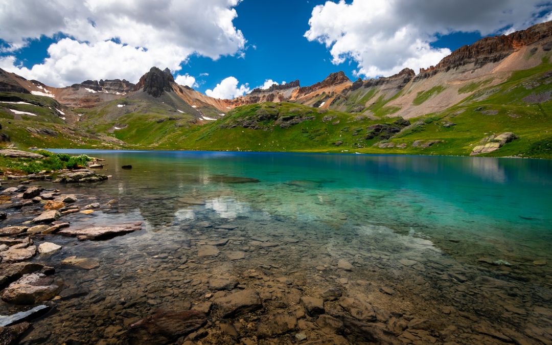 Crystal clear turquoise alpine lake reflecting rocky mountains under blue sky with white clouds and green slopes.