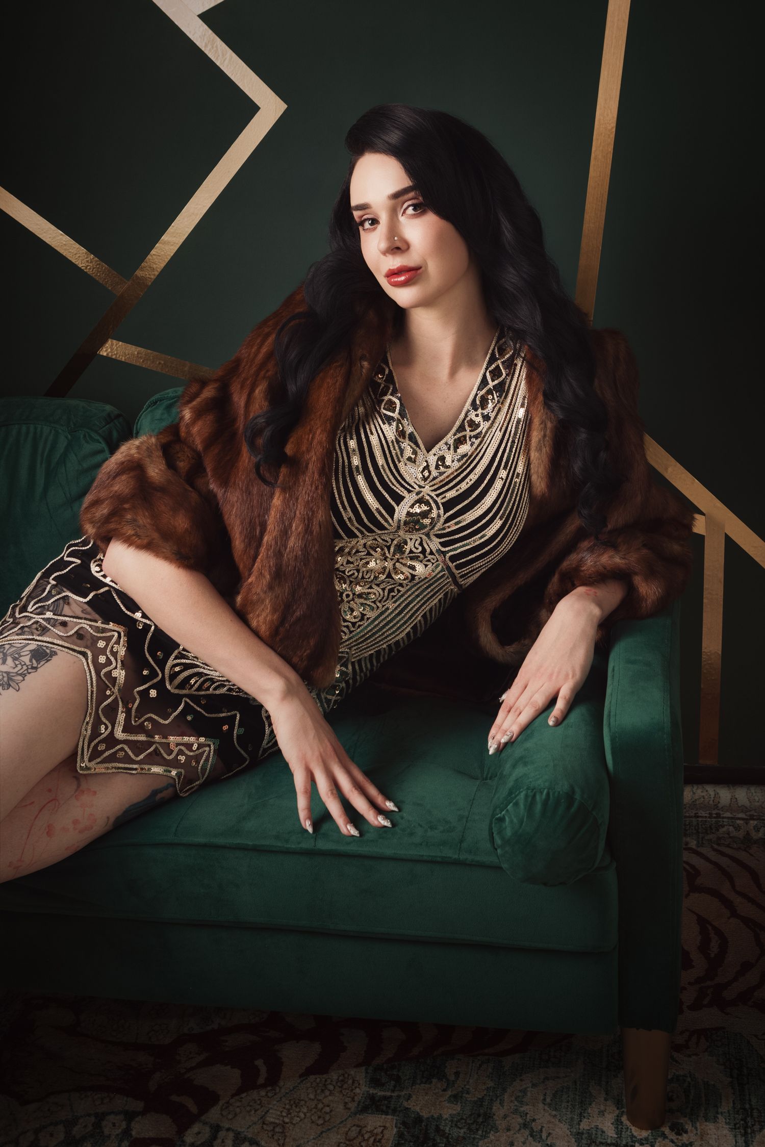 Elegant woman in gold embroidered dress and brown fur stole seated on green velvet chair against art deco wall.
