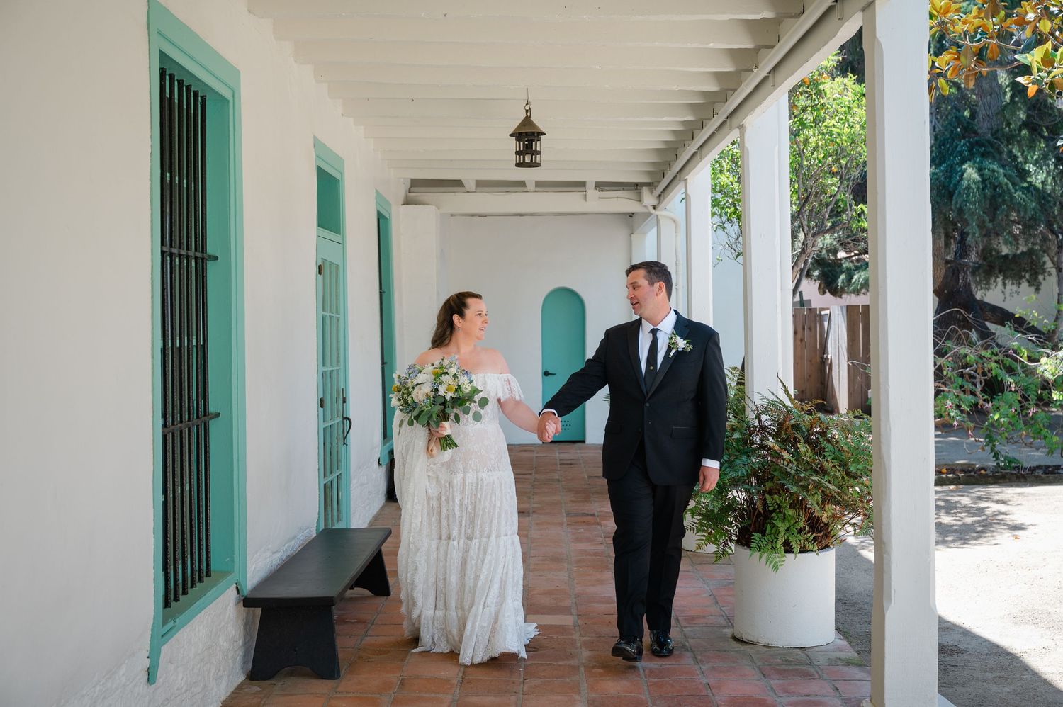 Bride and groom holding hands walking along a white colonial building with turquoise doors and terracotta tile floors.