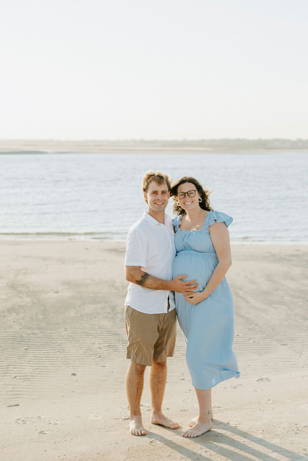Couple poses on sandy beach, pregnant woman in blue dress cradling bump with partner in white shirt and khaki shorts.