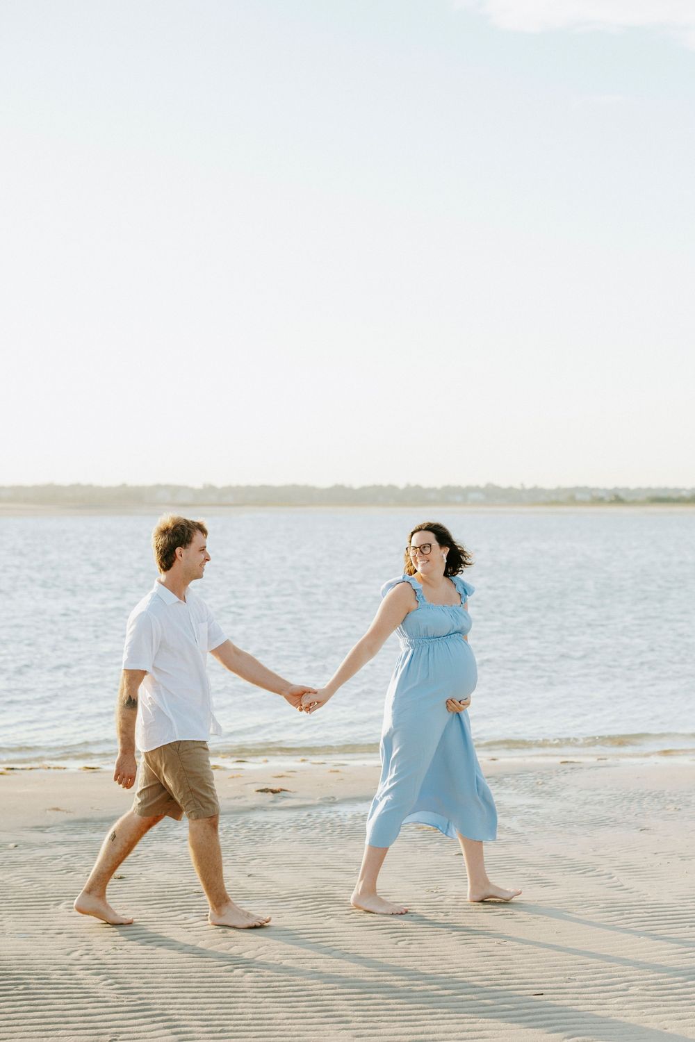 Pregnant woman in blue dress holds hands with man in white shirt walking barefoot on sandy beach by calm water.