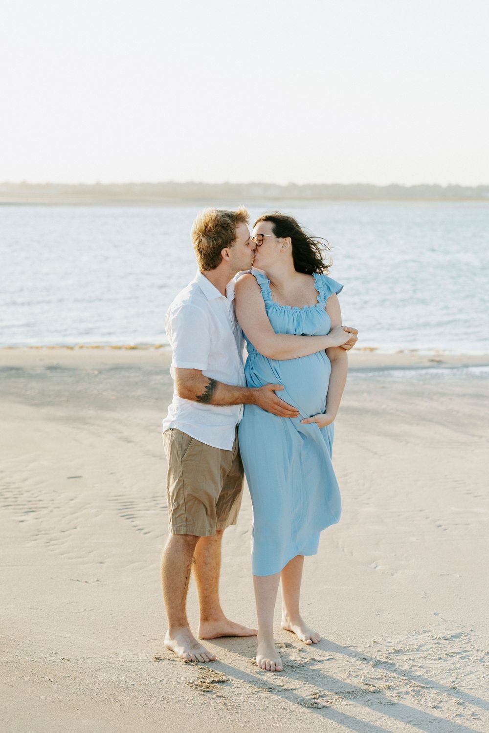 Couple sharing a kiss on a sandy beach, pregnant woman in a blue dress embraced by partner in white shirt.