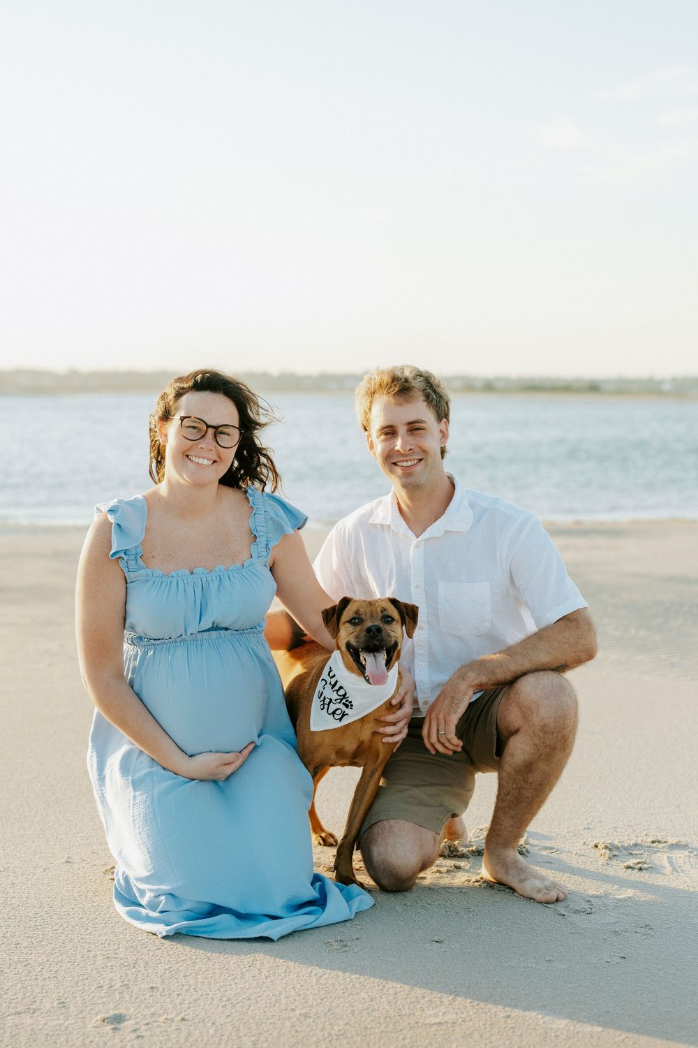 Pregnant woman in blue dress poses with husband and dog wearing bandana on sandy beach at sunset.