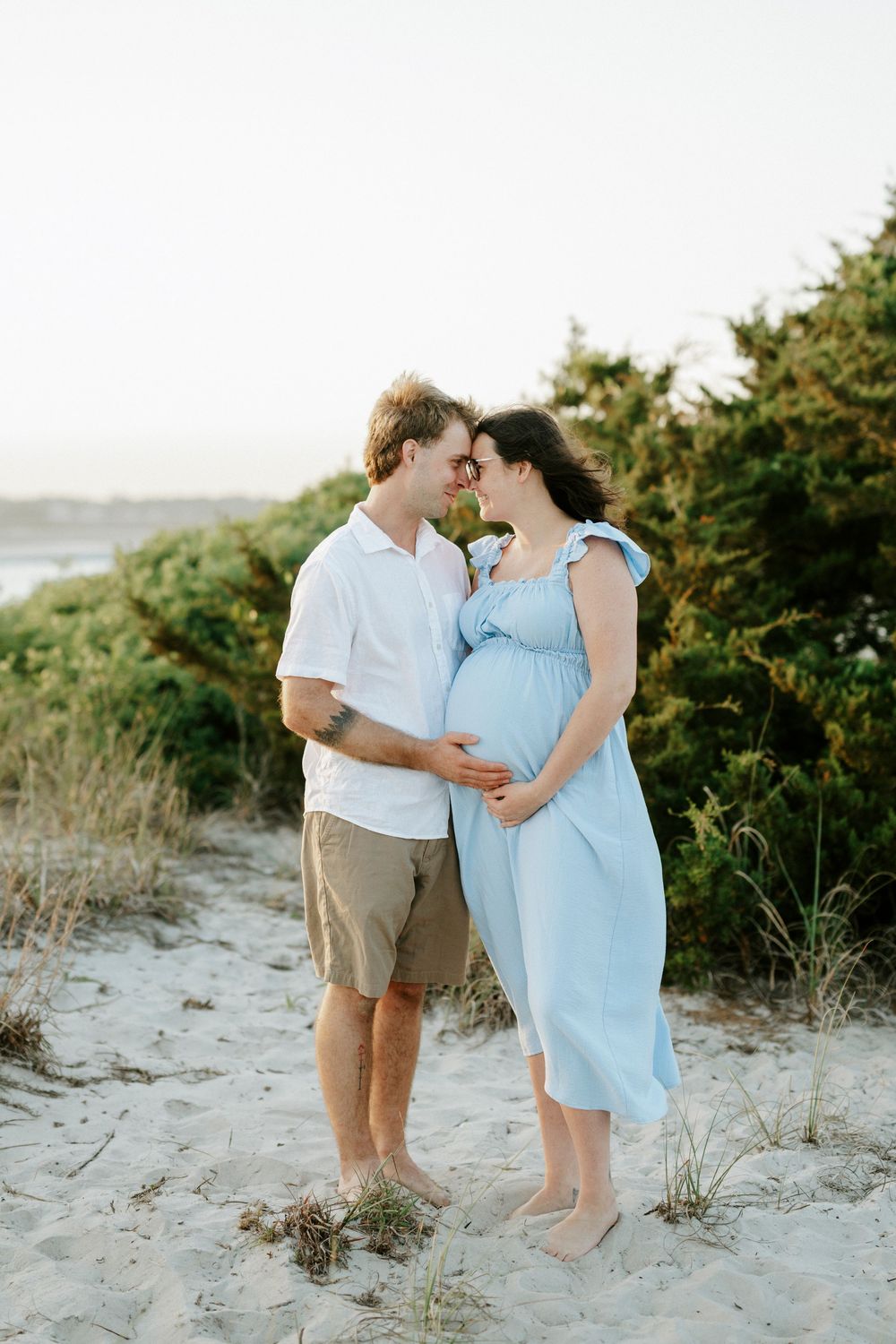 Pregnant couple embracing on sandy beach dunes, man in white shirt and woman in light blue maternity dress.