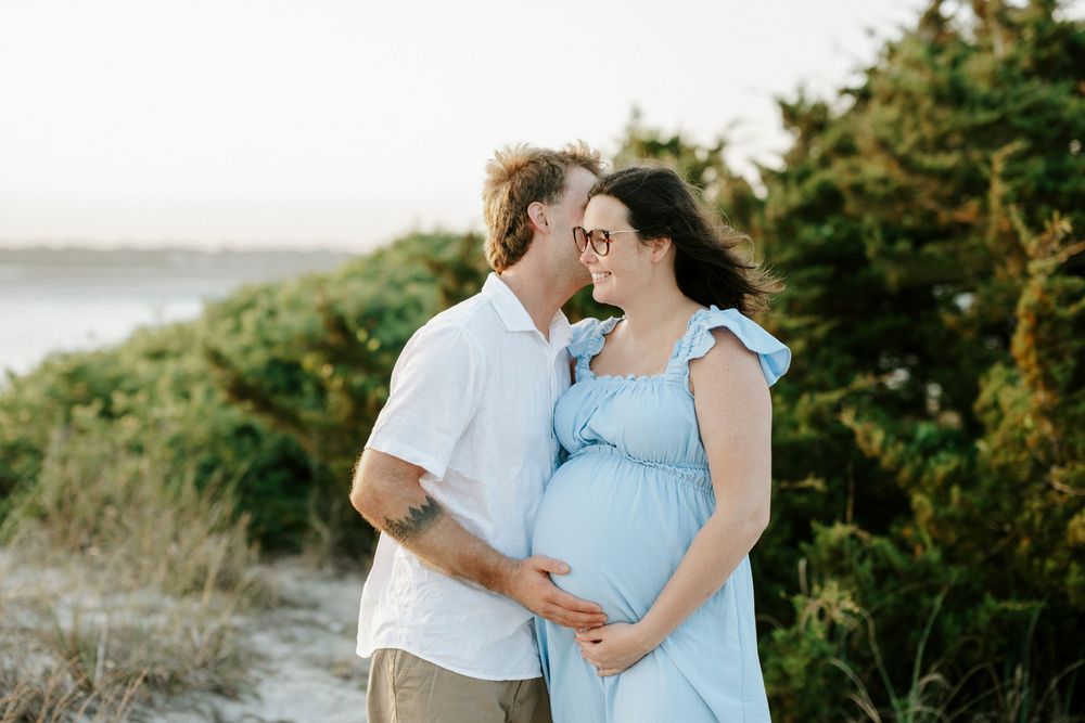 Couple sharing a tender moment on a sandy beach, woman in blue maternity dress, man in white shirt, green shrubs behind.