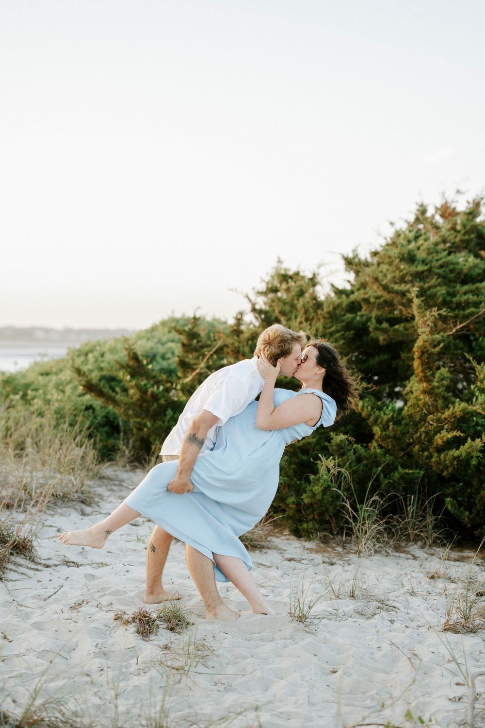 Couple shares romantic dip kiss on sandy beach dunes surrounded by lush green coastal shrubs.