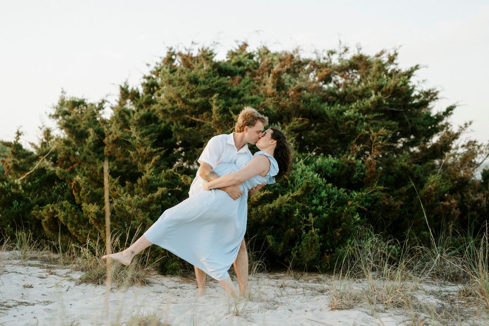 Man dipping pregnant woman in blue dress for a romantic kiss on sandy beach with green trees behind.