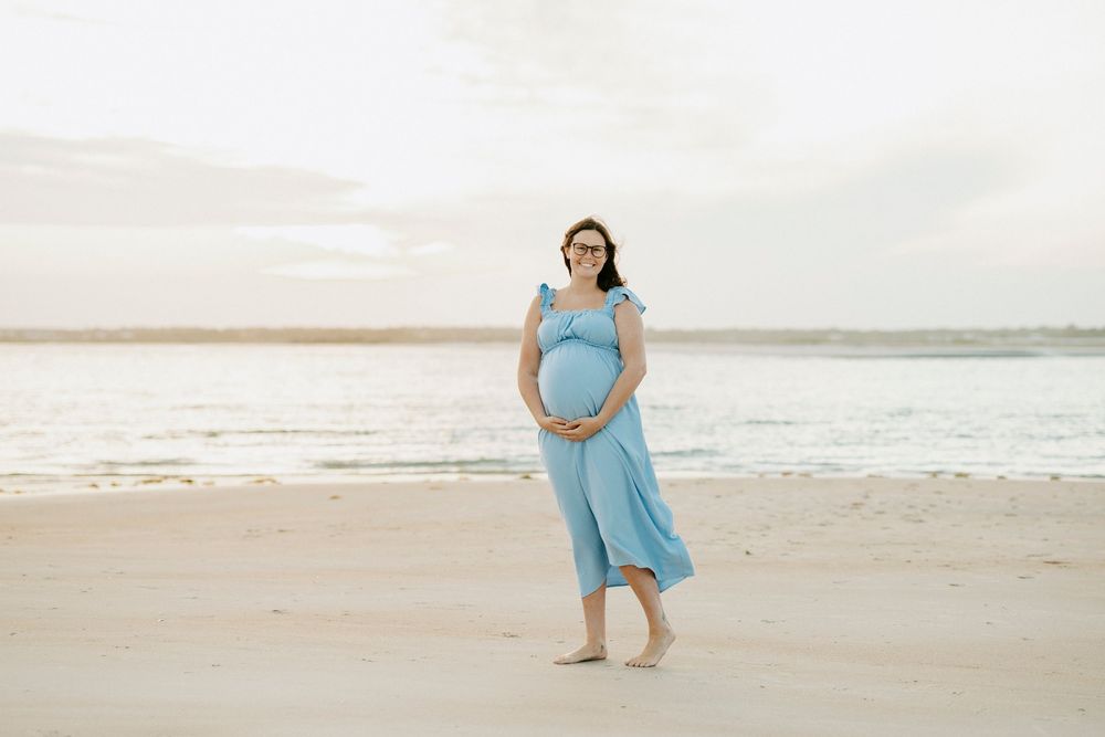 Pregnant woman in blue dress smiling on sandy beach, holding baby bump, ocean backdrop at sunset.