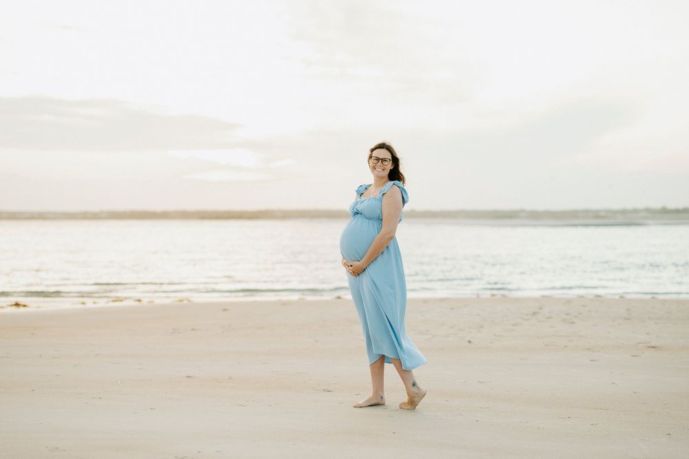 Pregnant woman in light blue dress walking on sandy beach near ocean during golden hour maternity photoshoot.