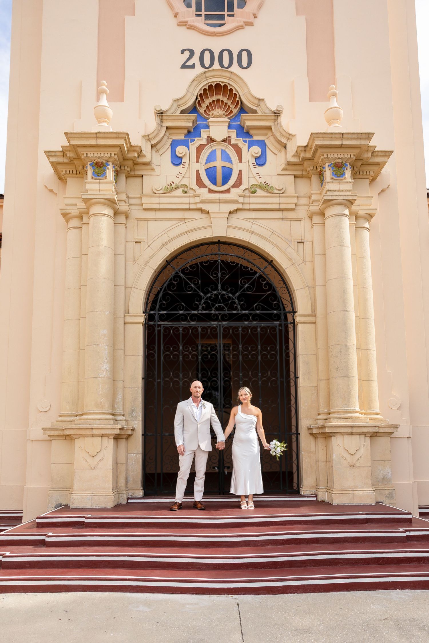 Bride and groom holding hands on steps of ornate baroque church entrance with number 2000 above doorway.