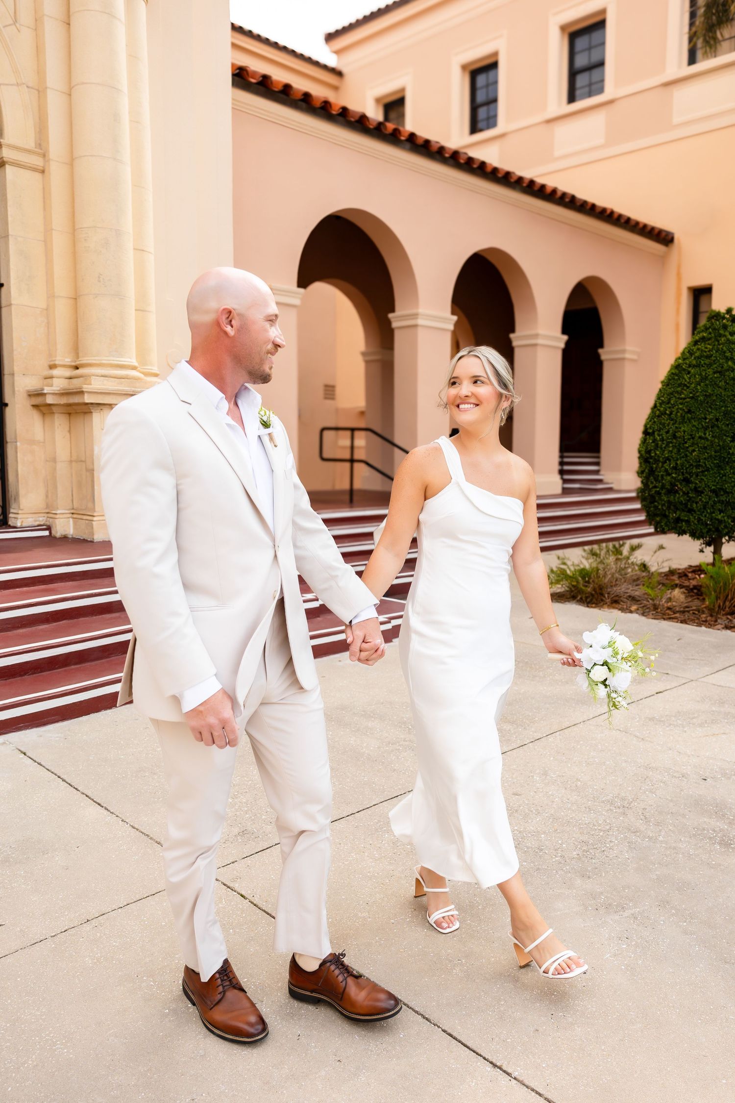 Bride and groom in white attire hold hands walking outside a Spanish-style building on their wedding day.
