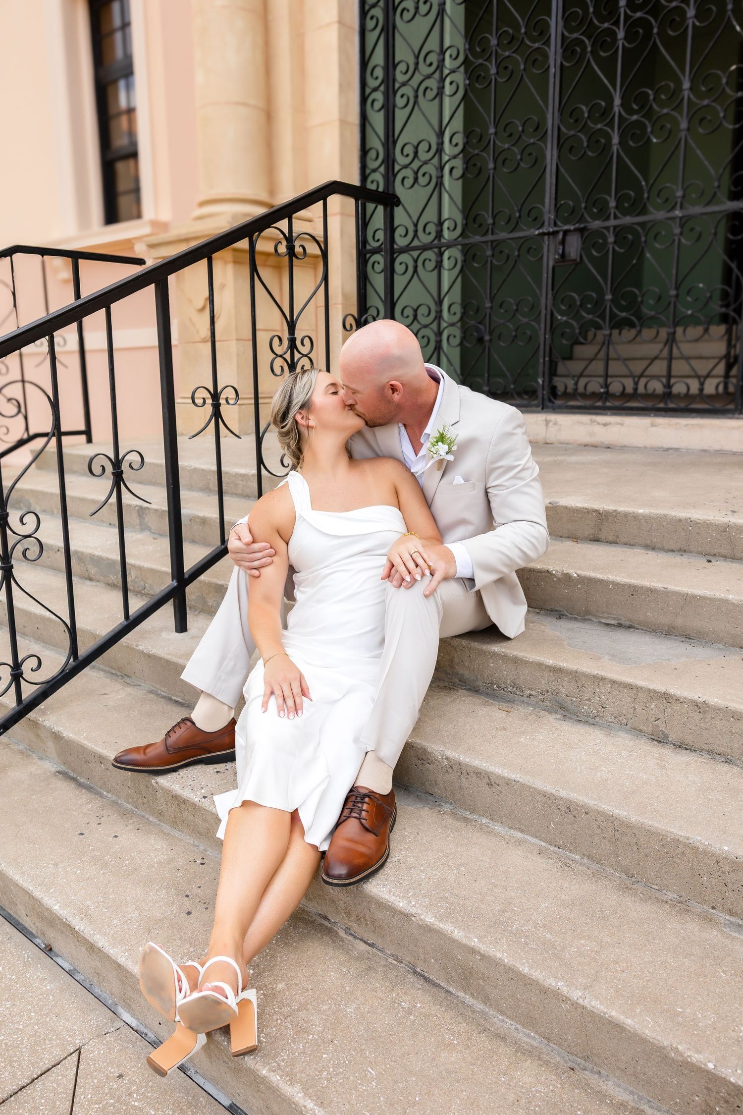 Bride and groom sitting on church steps sharing a kiss, wearing white dress and light gray suit with brown shoes.