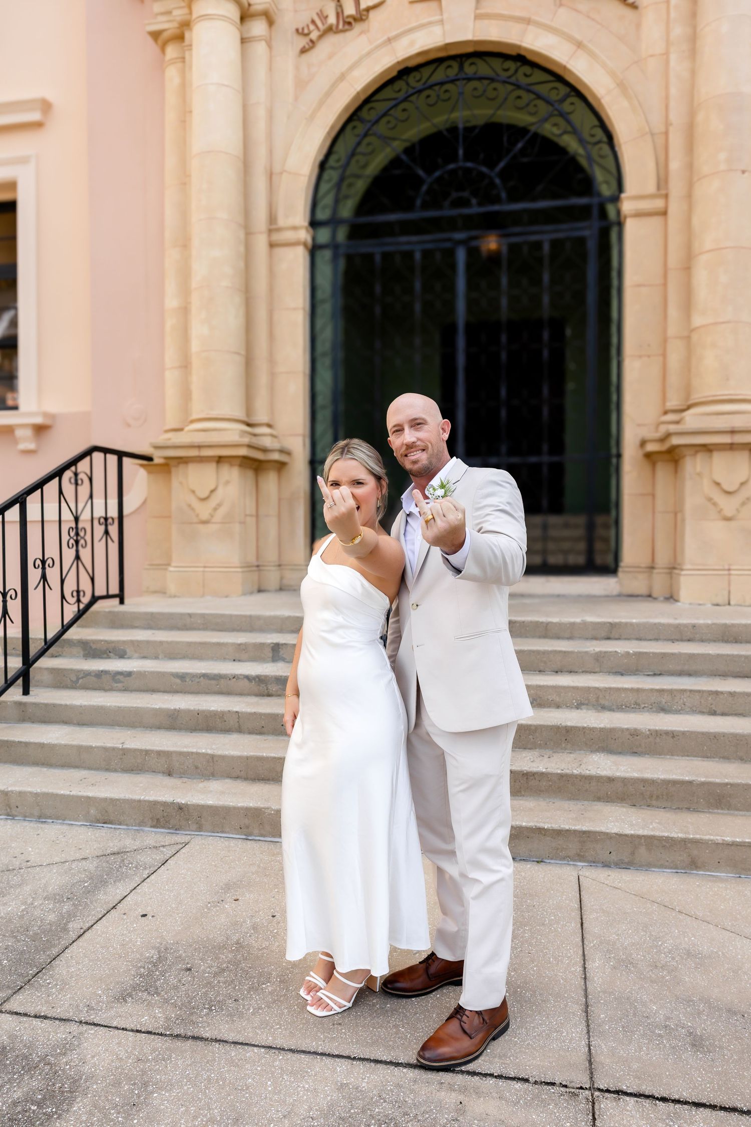 Newlywed couple showing off wedding rings outside elegant building, bride in white dress, groom in light suit.