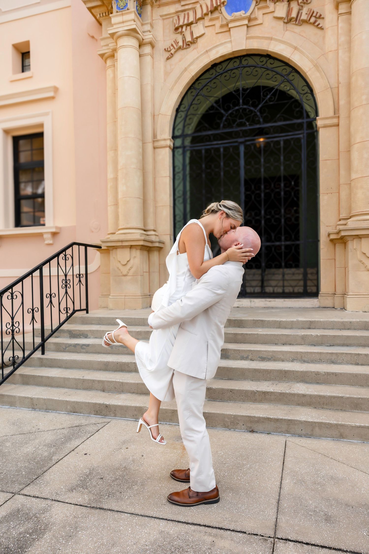 Couple in white wedding attire sharing a romantic kiss, groom lifting bride on ornate building steps.