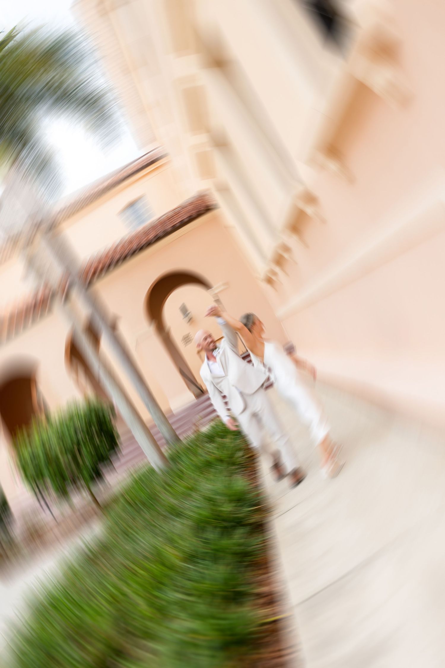 Blurred artistic zoom effect photo of a couple in wedding attire walking outside a Mediterranean-style venue.