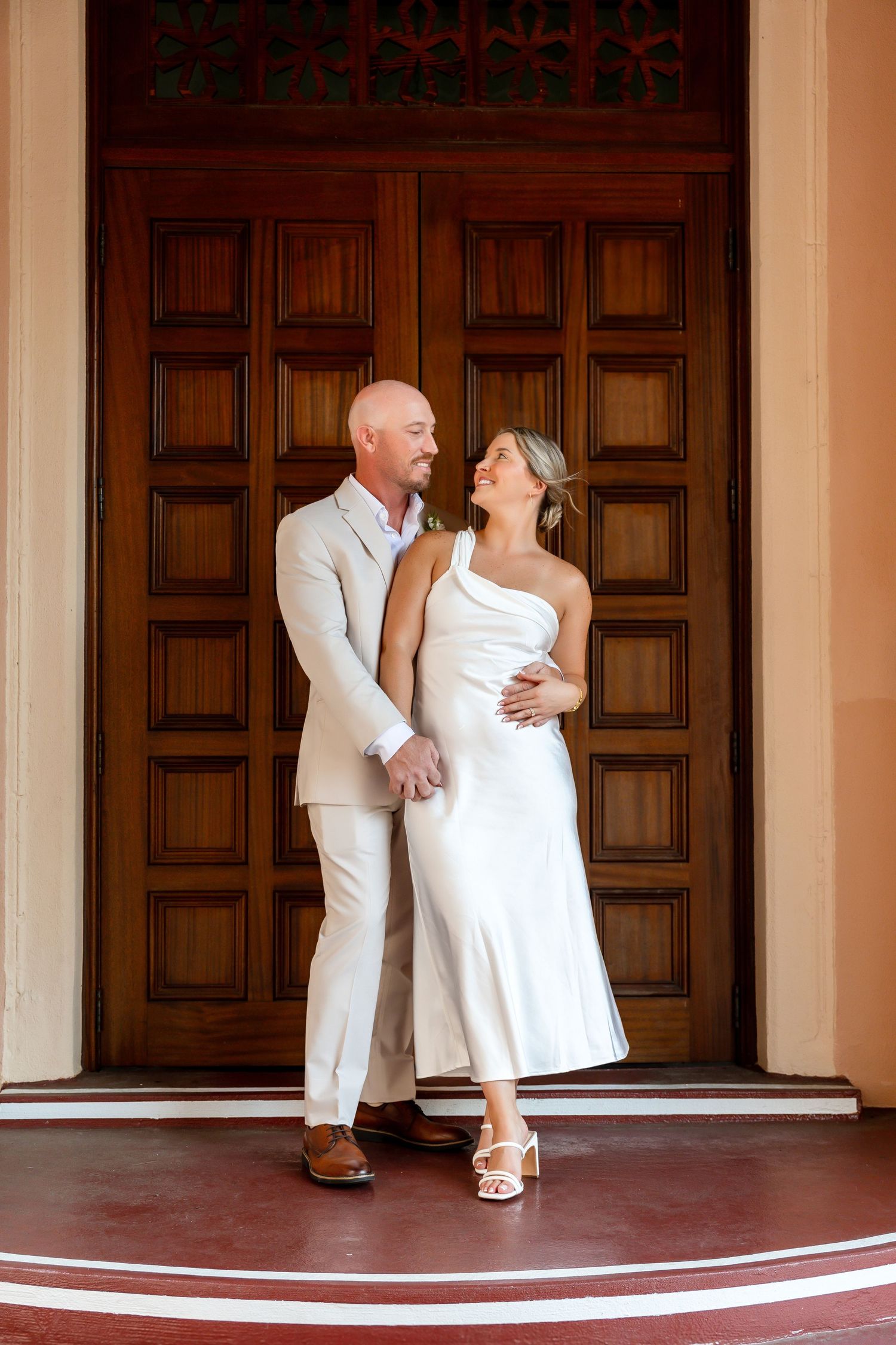 Bride in white dress and groom in beige suit pose together in front of elegant wooden doors.