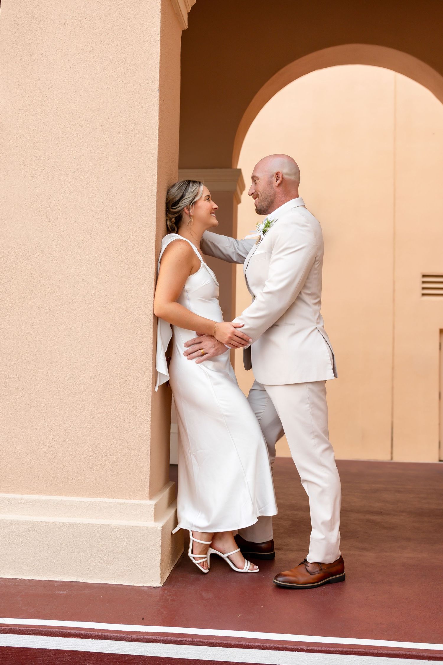 Bride and groom share a romantic moment under an arch, dressed in white wedding attire.