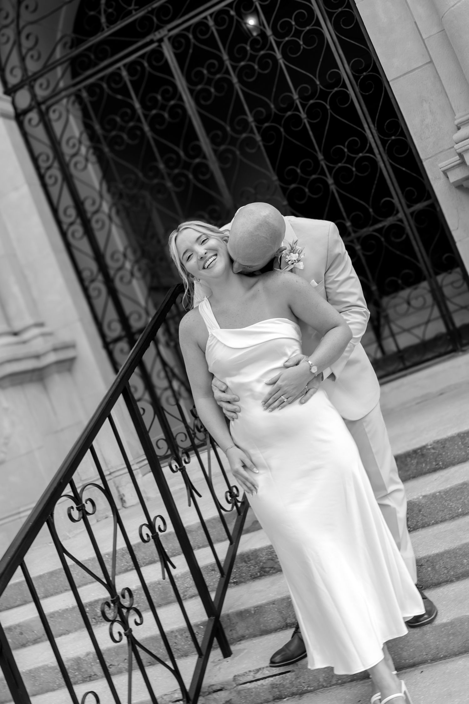 Black and white photo of a smiling bride in white dress embraced by groom on elegant stone steps.