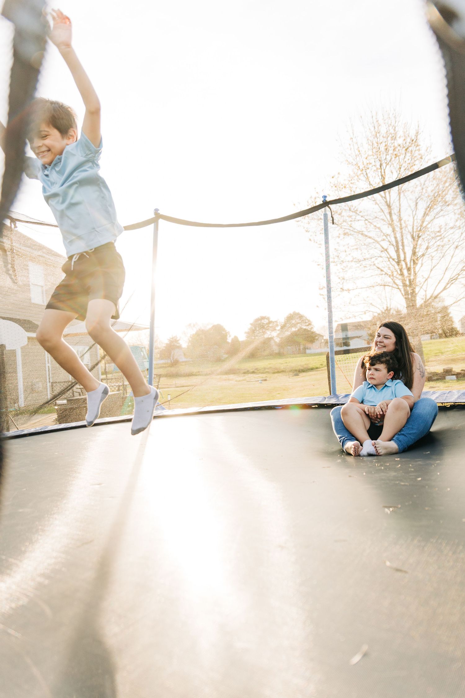 A boy jumping on a trampoline while a woman and young child sit and watch in a sunny backyard.