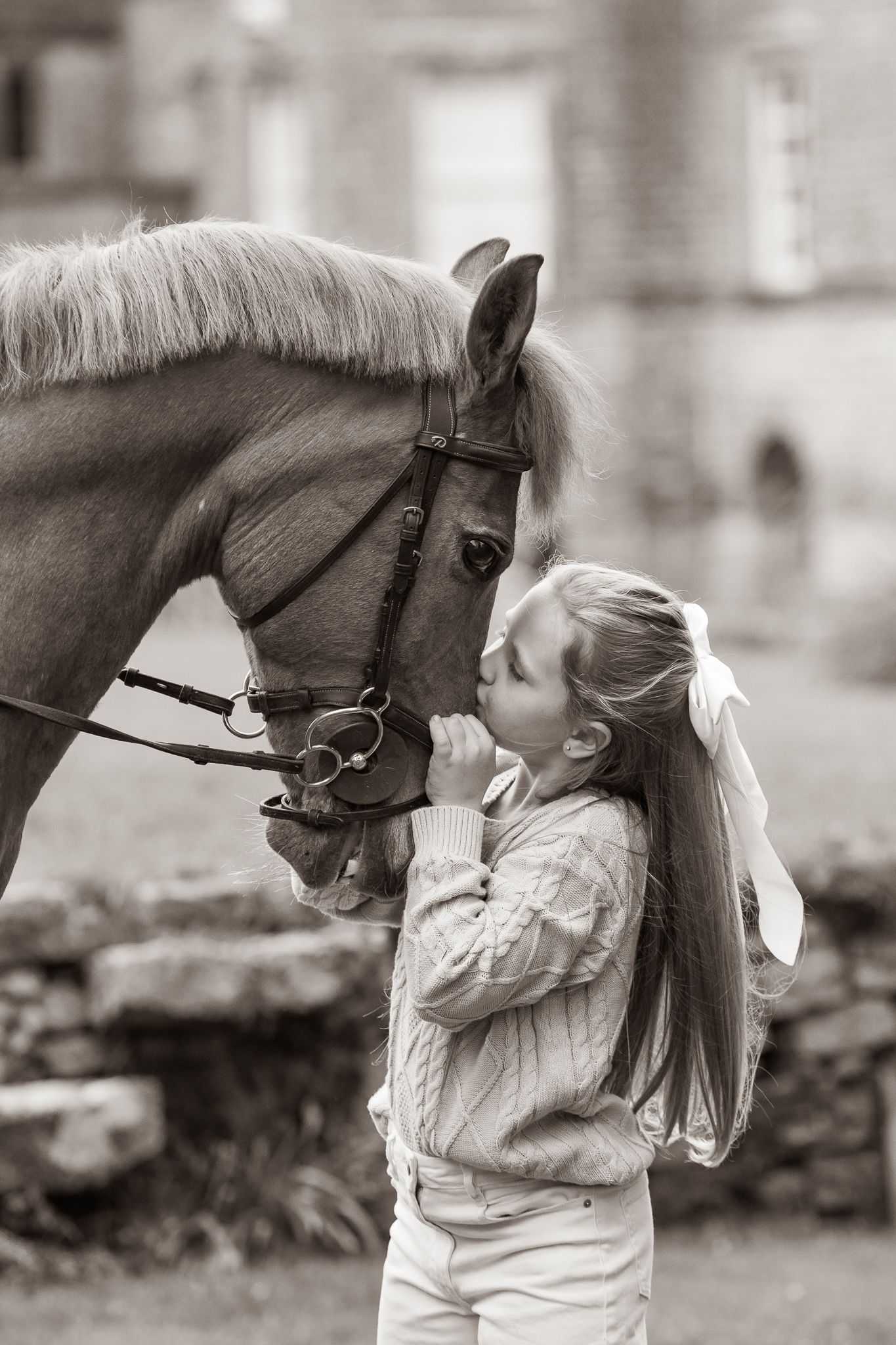 Black and white photo of a young girl with long hair kissing a brown horse wearing a bridle outdoors.