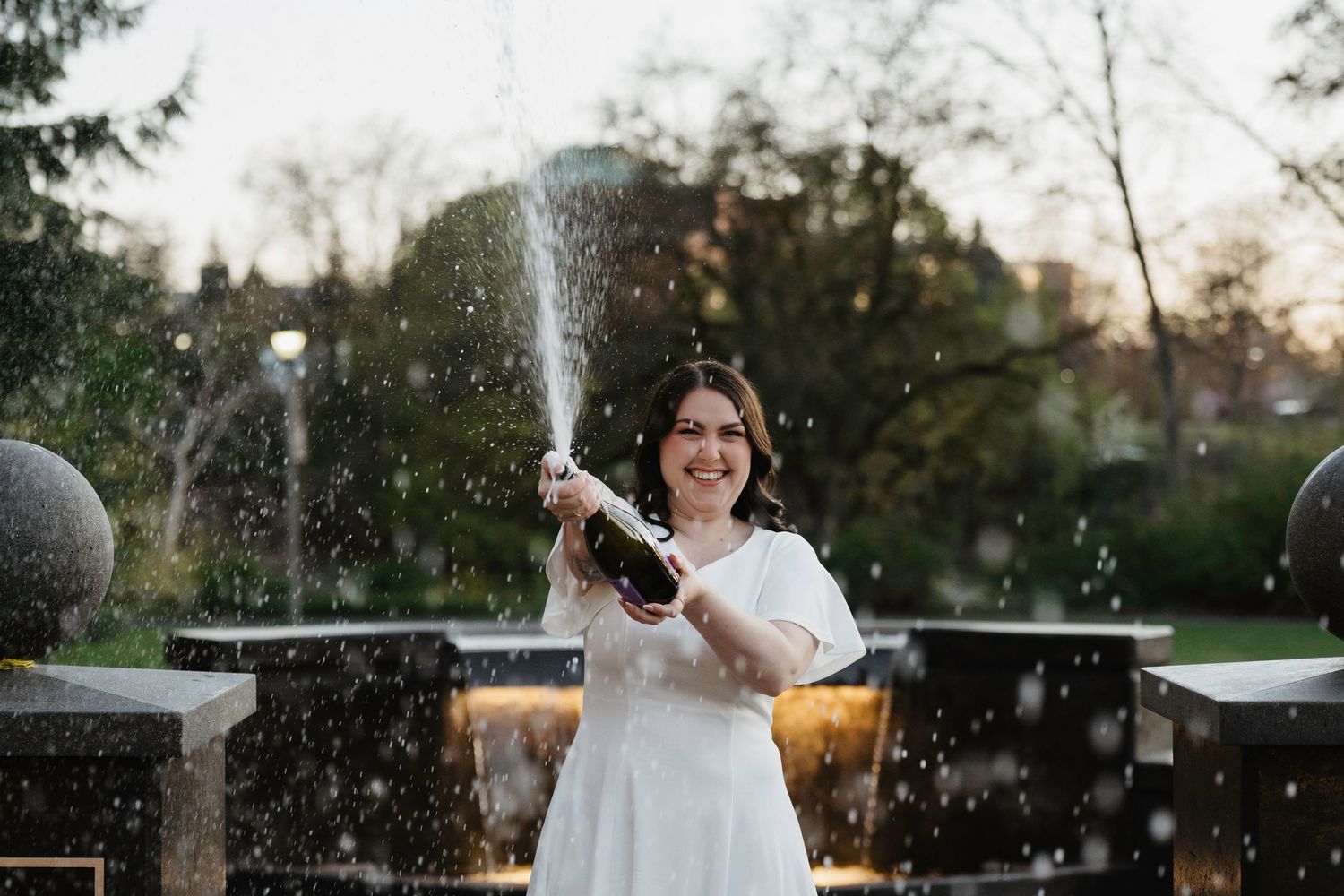 Woman in white dress joyfully popping champagne bottle outdoors near fountain in lush green park setting.