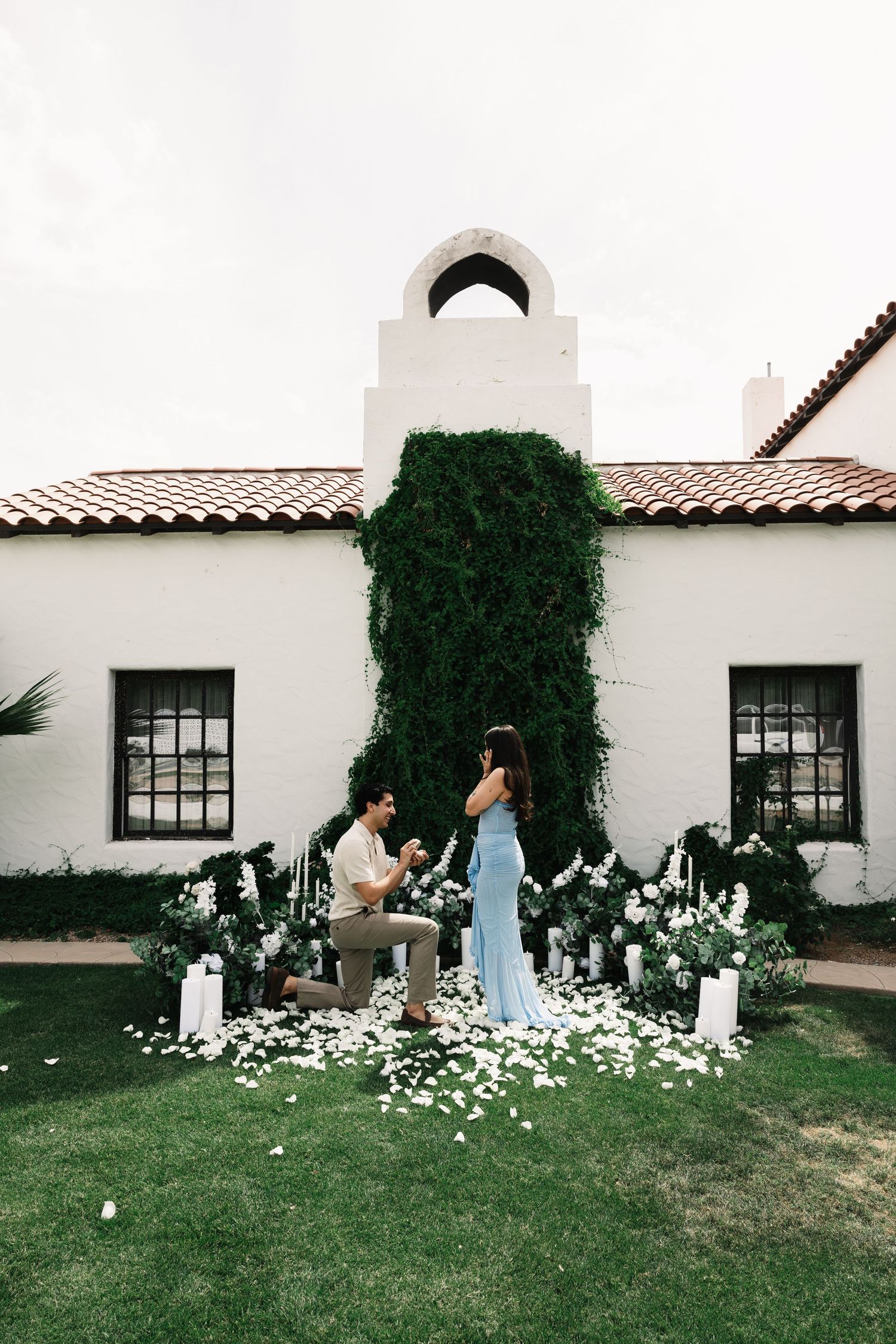 Man kneeling proposing to woman in blue dress outdoors, surrounded by white flowers and candles near white Spanish-style building.