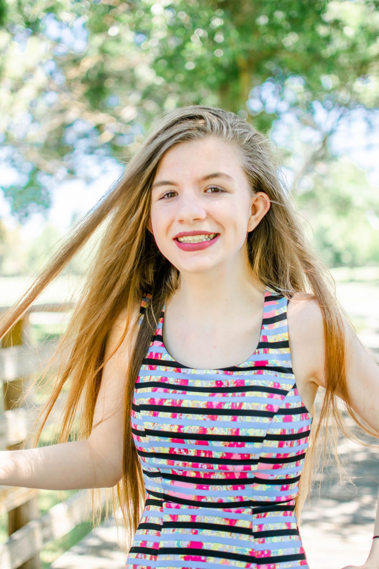 Smiling teenage girl with long blonde hair and braces wearing a colorful striped dress outdoors in a sunny park.