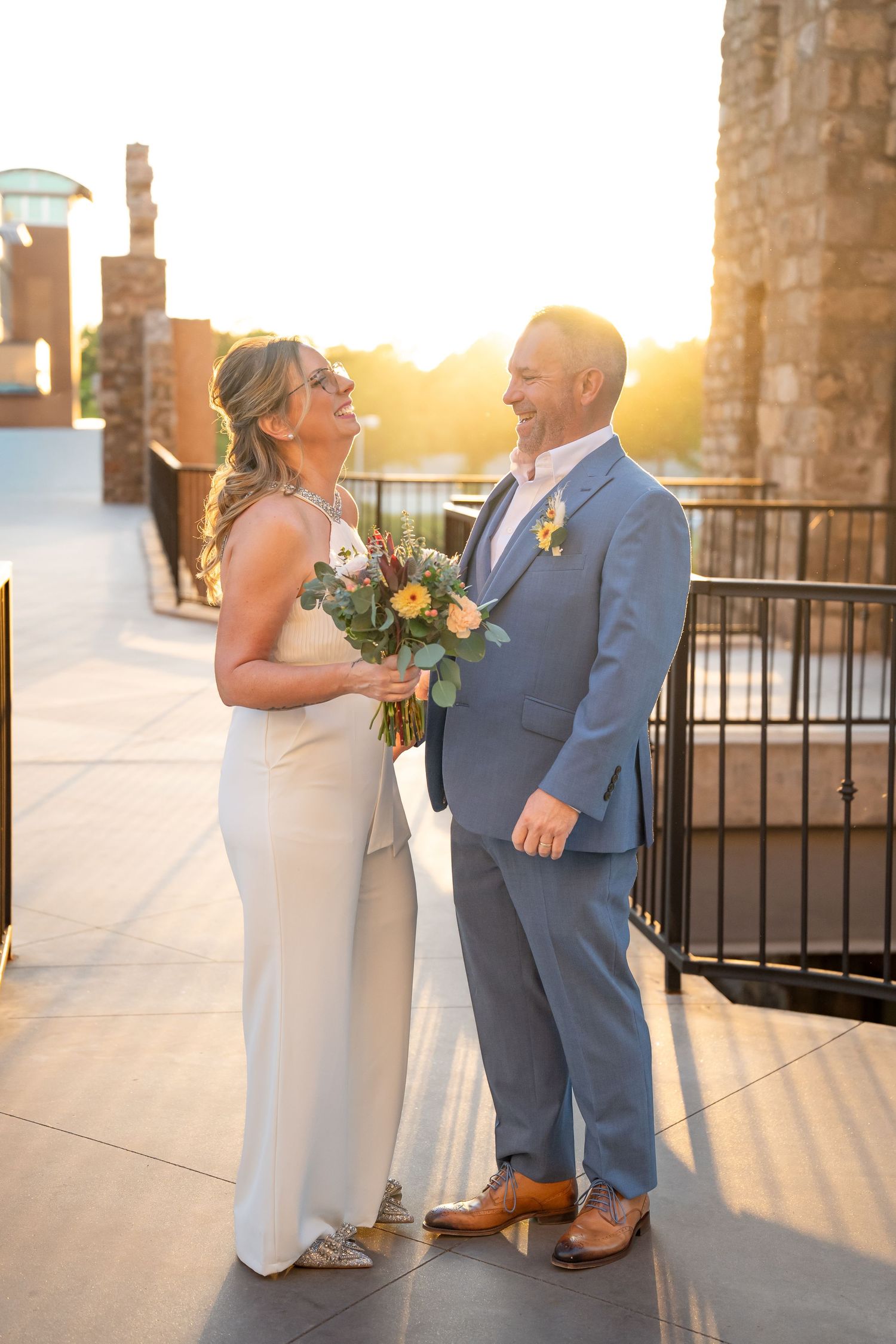 Bride and groom laughing joyfully together outdoors at golden hour, bride holding a pastel bouquet in wedding attire.