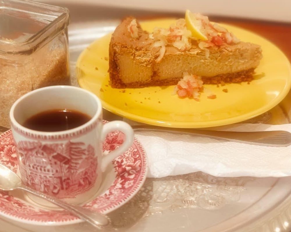 A slice of lemon cheesecake on a yellow plate served with a cup of black coffee in a red and white mug.