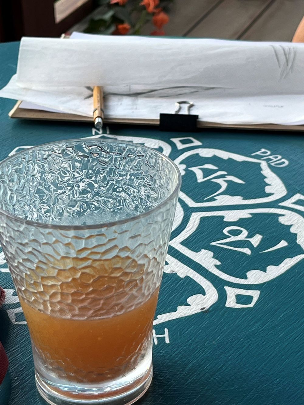 A textured glass of amber cocktail on a teal patterned table mat with a menu and pencil in background.
