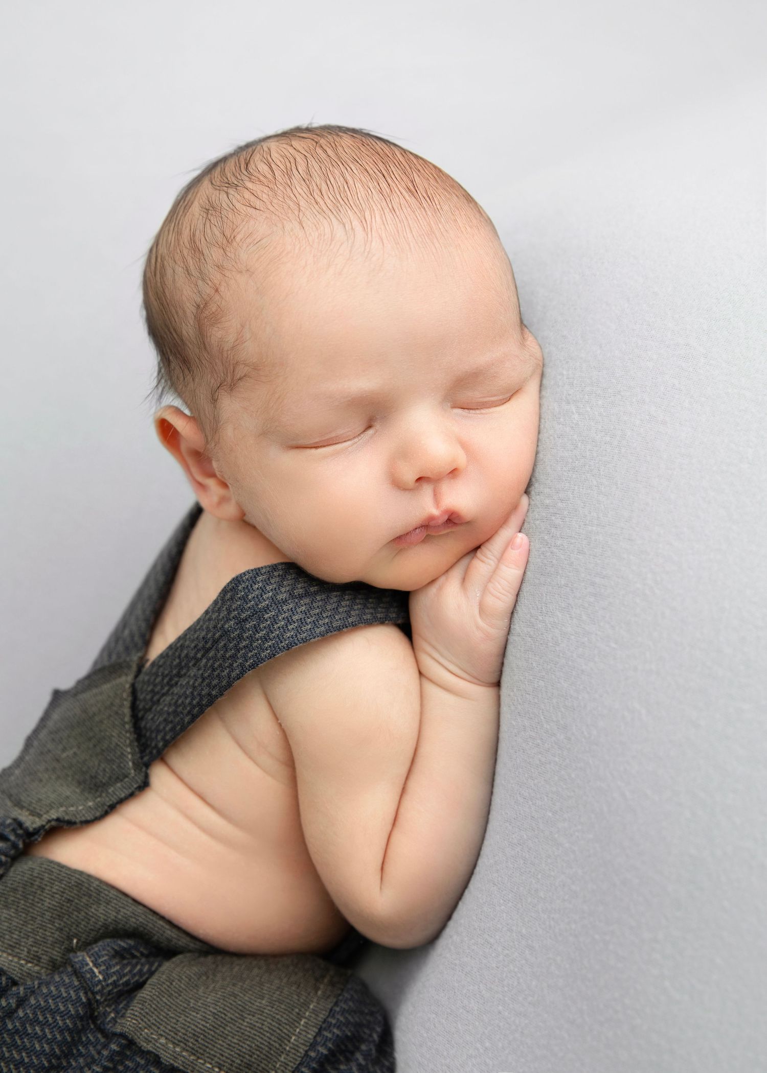 Peaceful newborn baby sleeping in gray overalls, resting cheek on tiny hands against a soft light gray background.