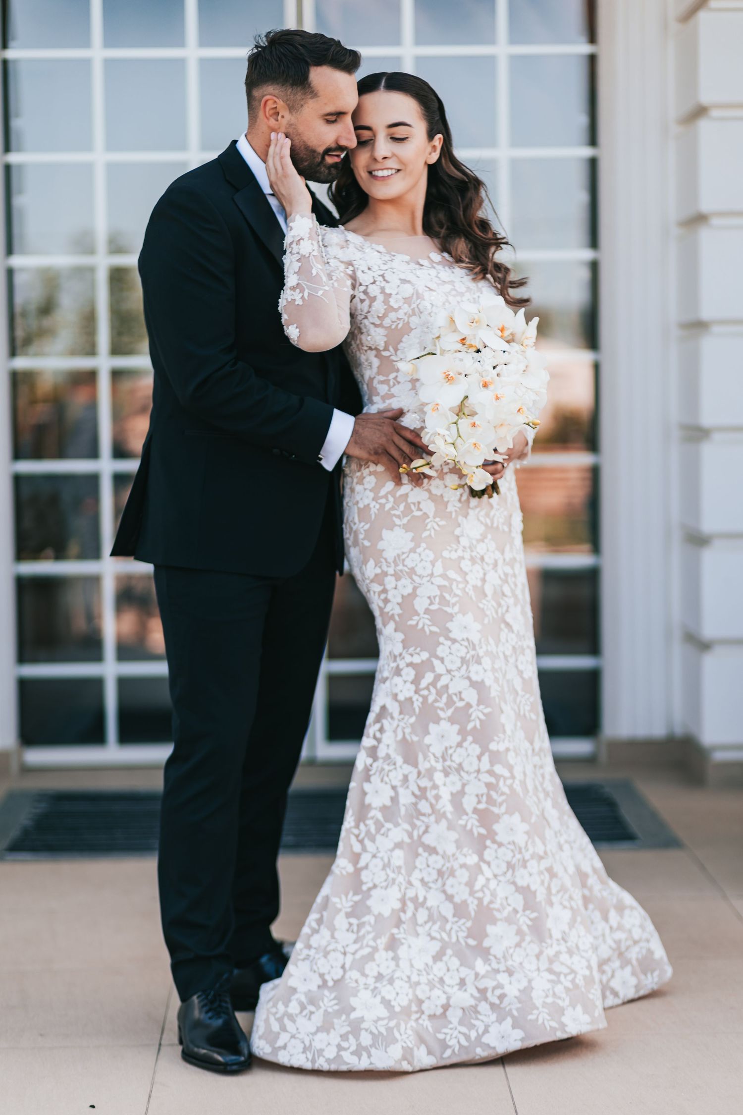 A bride in lace gown holds white bouquet as groom in black suit embraces her outside elegant venue doors.
