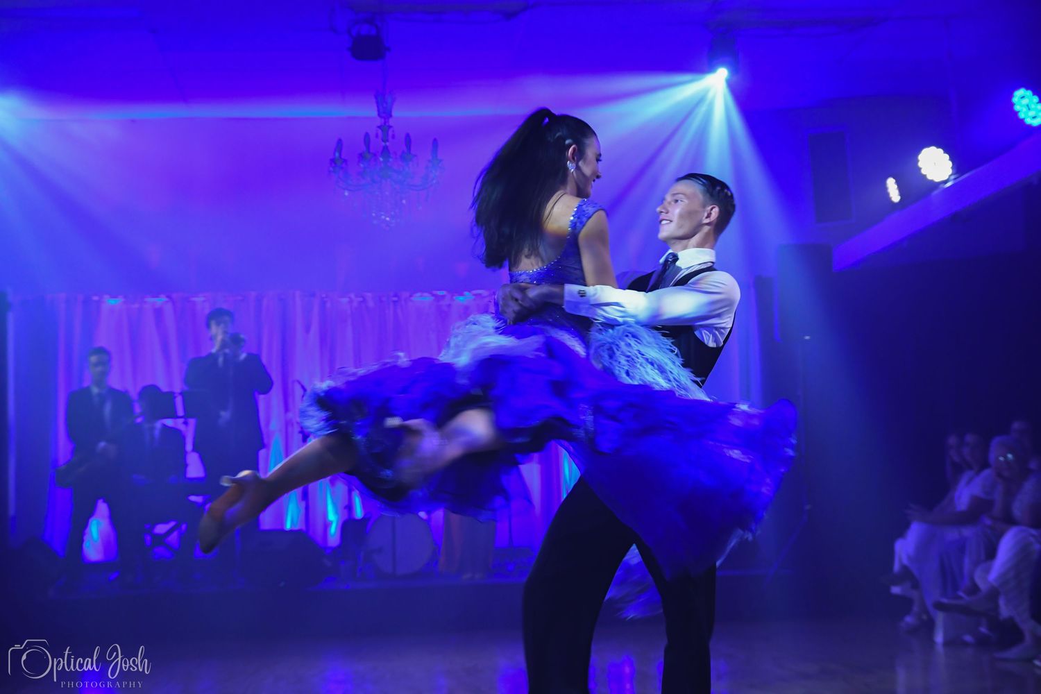 Dancers perform dramatic lift during ballroom dance under blue stage lighting.