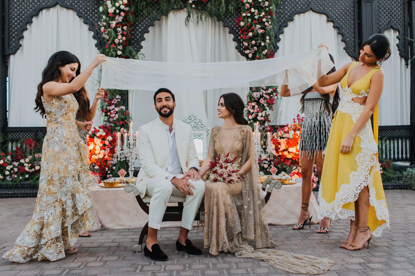 A traditional Indian wedding party poses together in front of a floral arch, wearing elegant attire in gold and yellow tones.