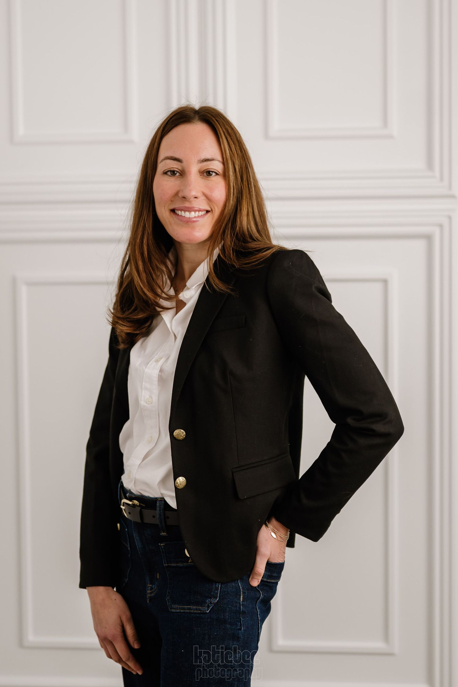 Professional wearing a classic black blazer with gold buttons, white button-up shirt and dark jeans against a white wall.