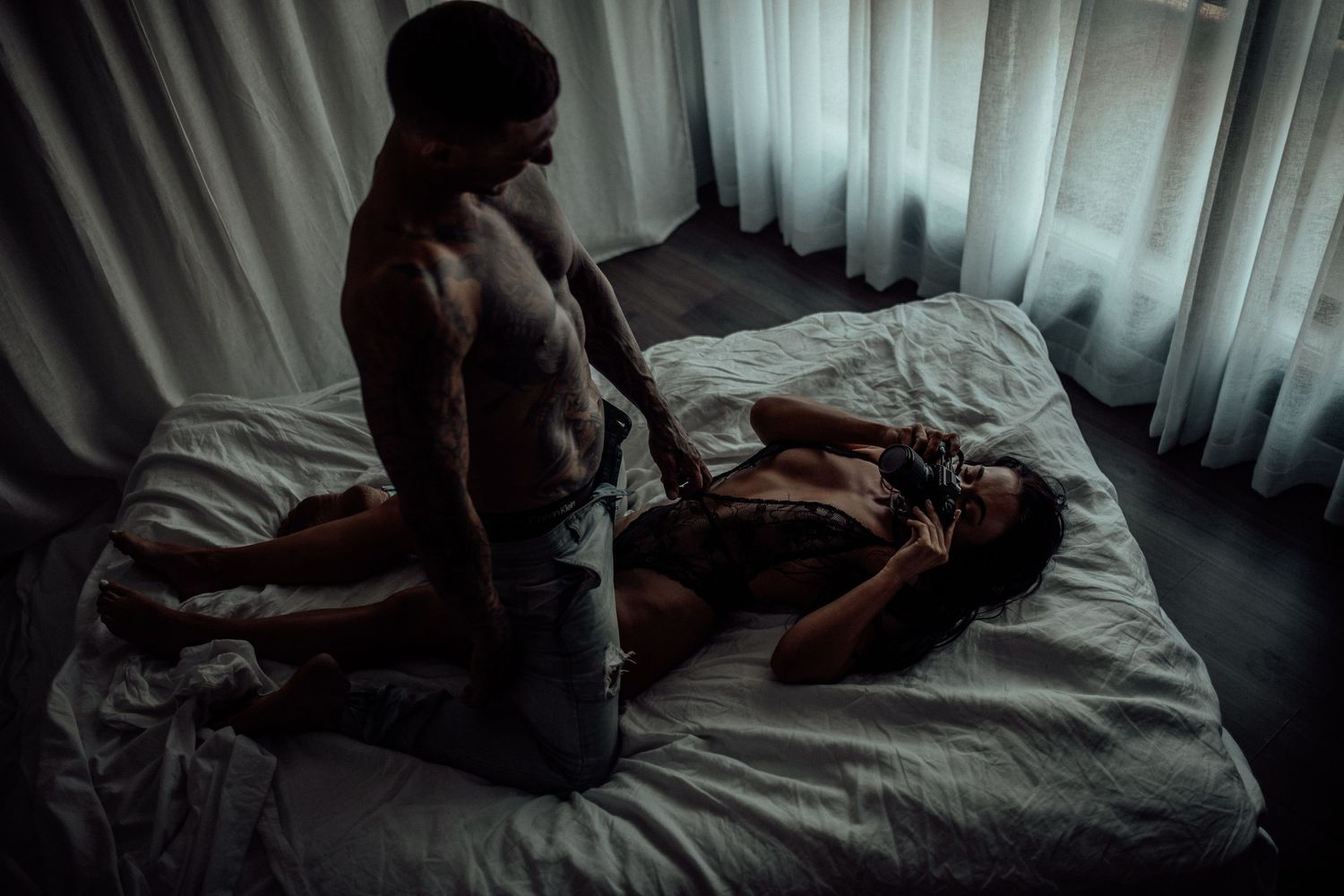 A dramatic bedroom scene with moody lighting and two figures on a bed silhouetted against sheer curtains.