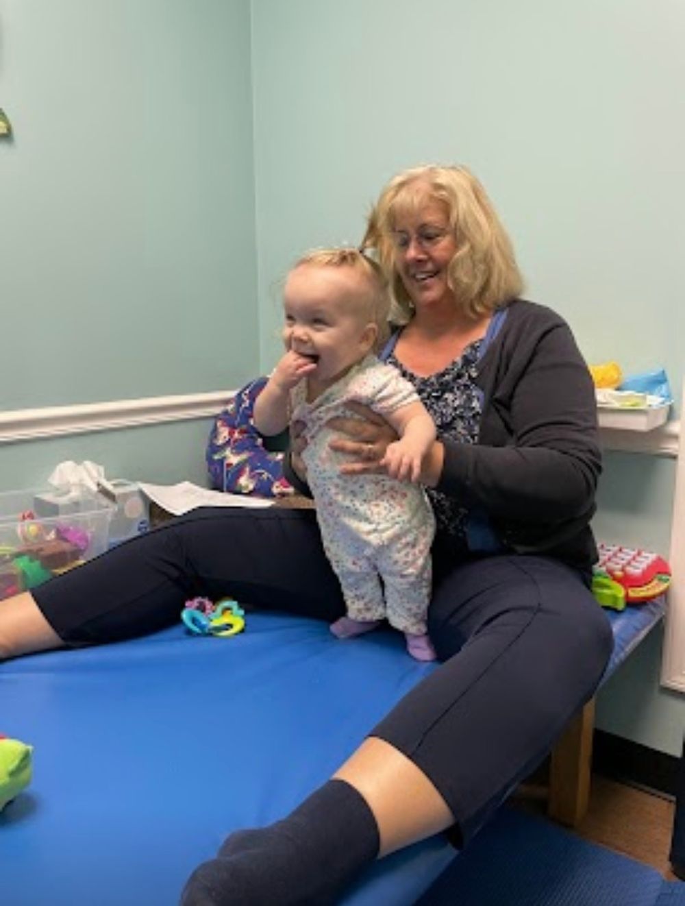 A person and toddler share a playful moment on a blue exam table during what appears to be a medical visit.