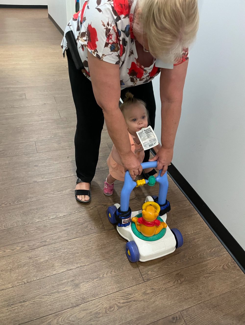 Someone helps a small child push a colorful toy lawnmower on a hardwood floor in an indoor setting.